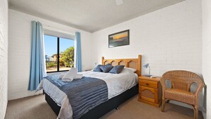 A bedroom with a bed, a nightstand, a wicker chair, a window with curtains, and a painting on the wall. - Apollo Unit 17 - Ground Floor (Narooma)