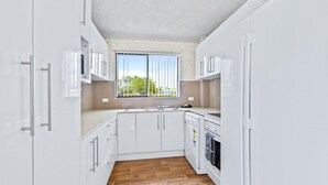 A modern kitchen with white cabinets, a wooden floor, and a window with vertical blinds. - Apollo Unit 20 - Ground Floor (Narooma)