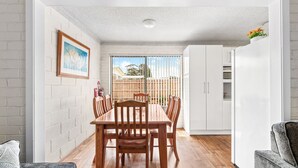 A dining area with a wooden table and chairs, white cabinets, and a view of a backyard with a wooden fence. - Apollo Unit 20 - Ground Floor (Narooma)