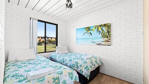 Ein Schlafzimmer mit zwei Betten, einem großen Fenster mit Blick auf ein Gebäude und einem Gemälde einer Strandansicht an der Wand.