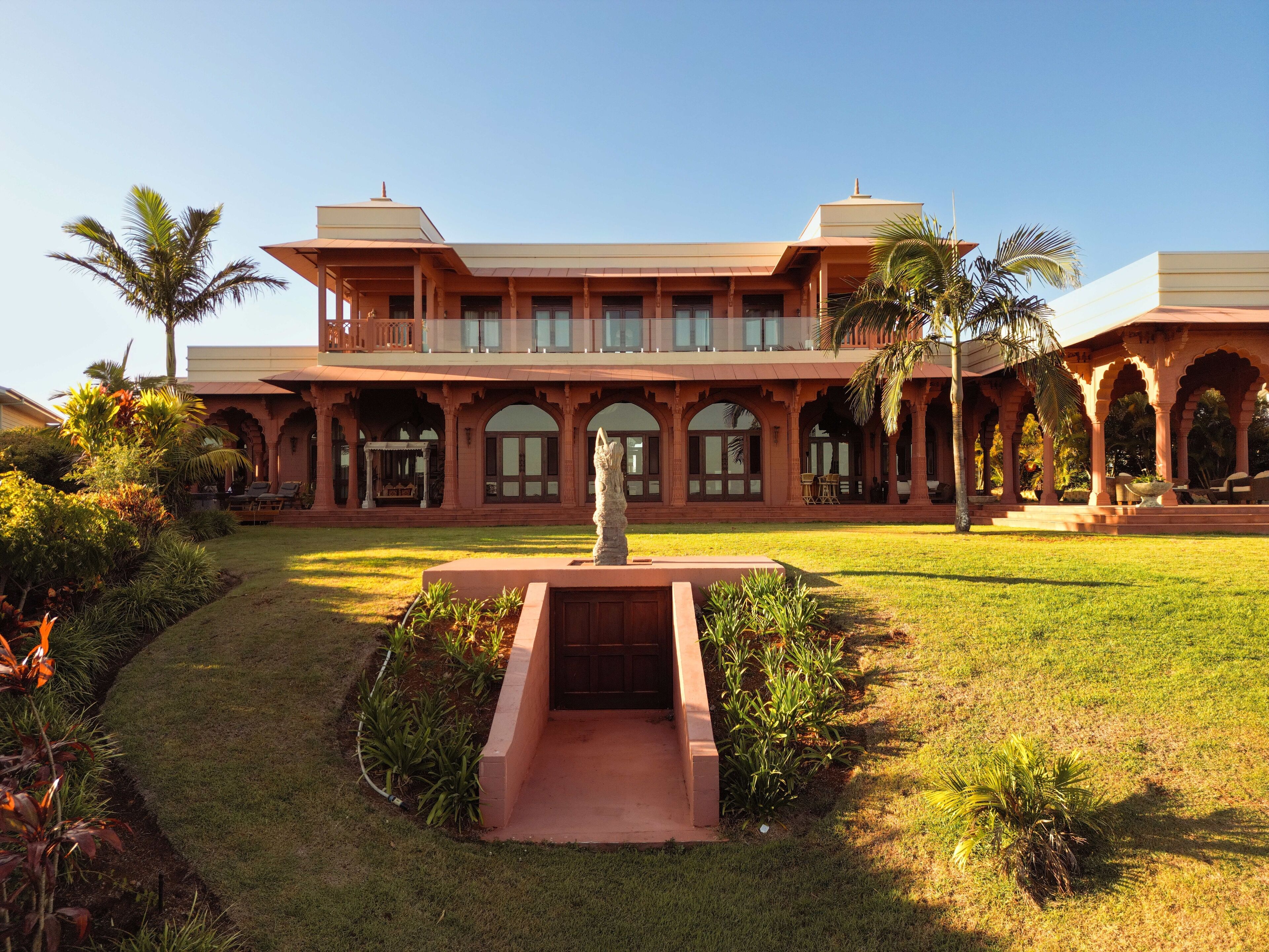 A large, two-story building with a central courtyard, featuring a statue, palm trees, and a well-maintained lawn.