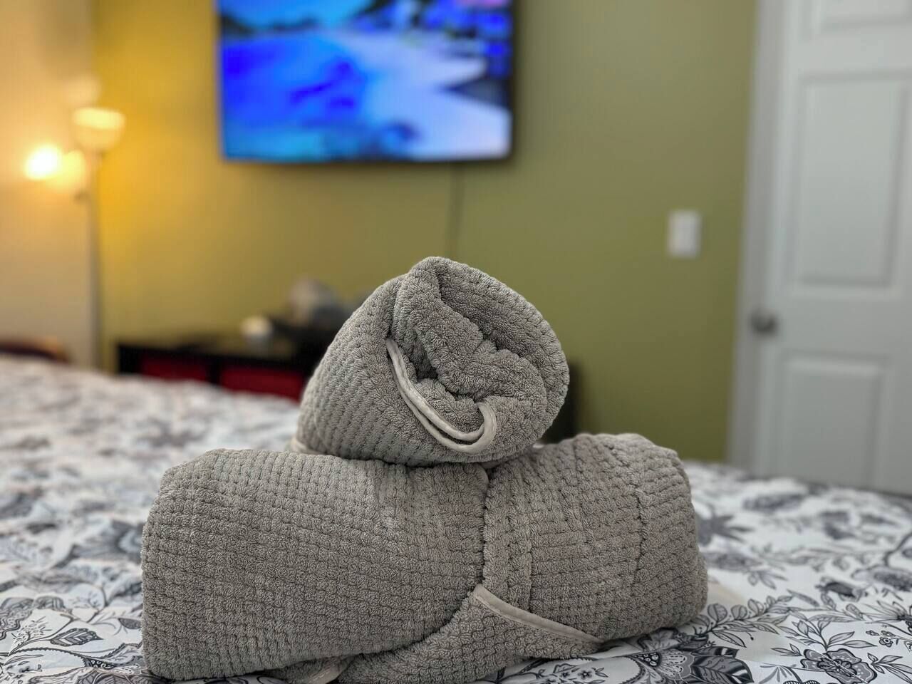 Ein ordentlich gefaltetes graues Handtuch auf einem Bett mit einer gemusterten Tagesdecke, ein Fernseher im Hintergrund und eine Tür zur rechten Seite.