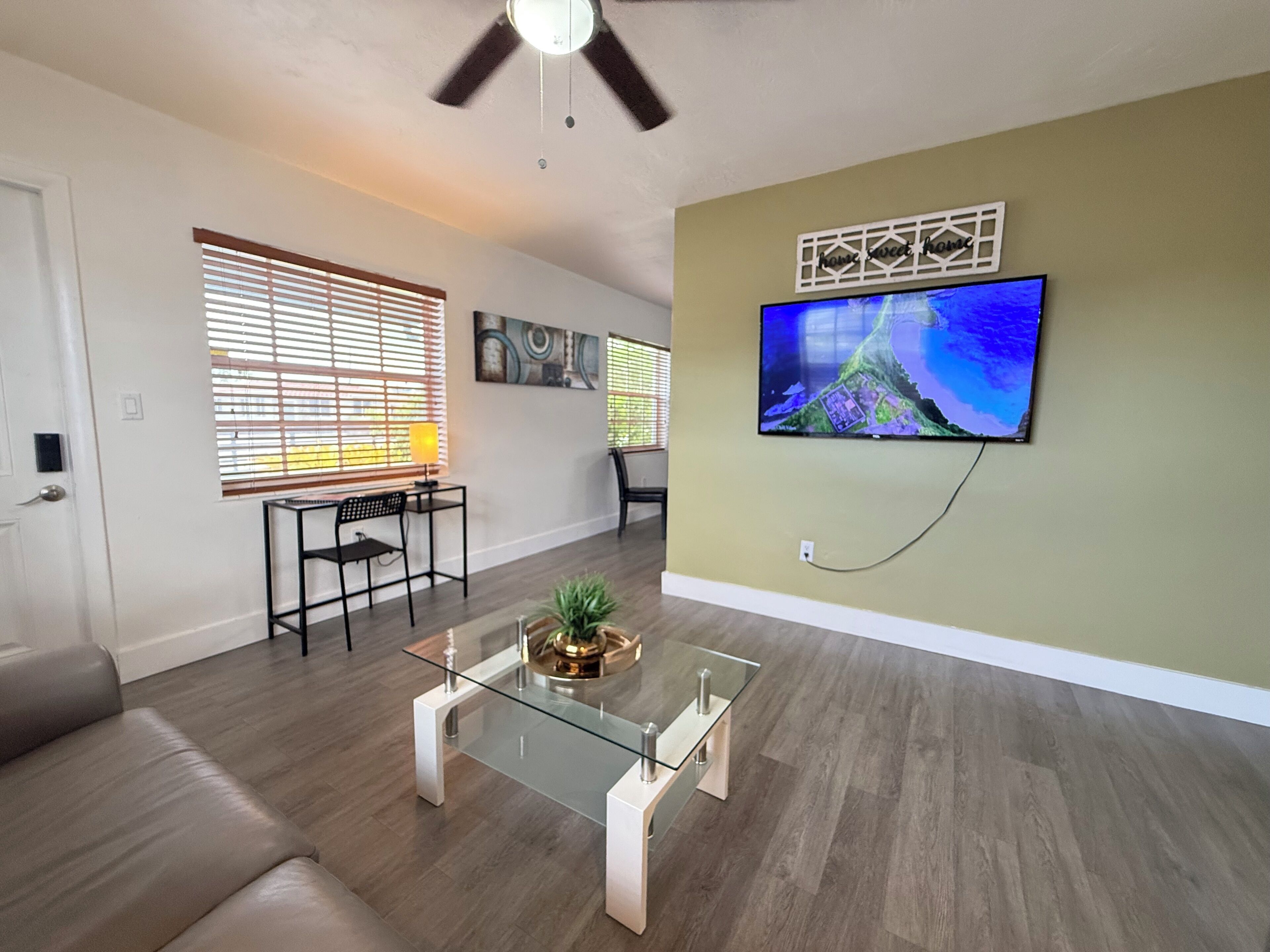 A living room with a sofa, a glass coffee table, a TV mounted on the wall, and a ceiling fan.