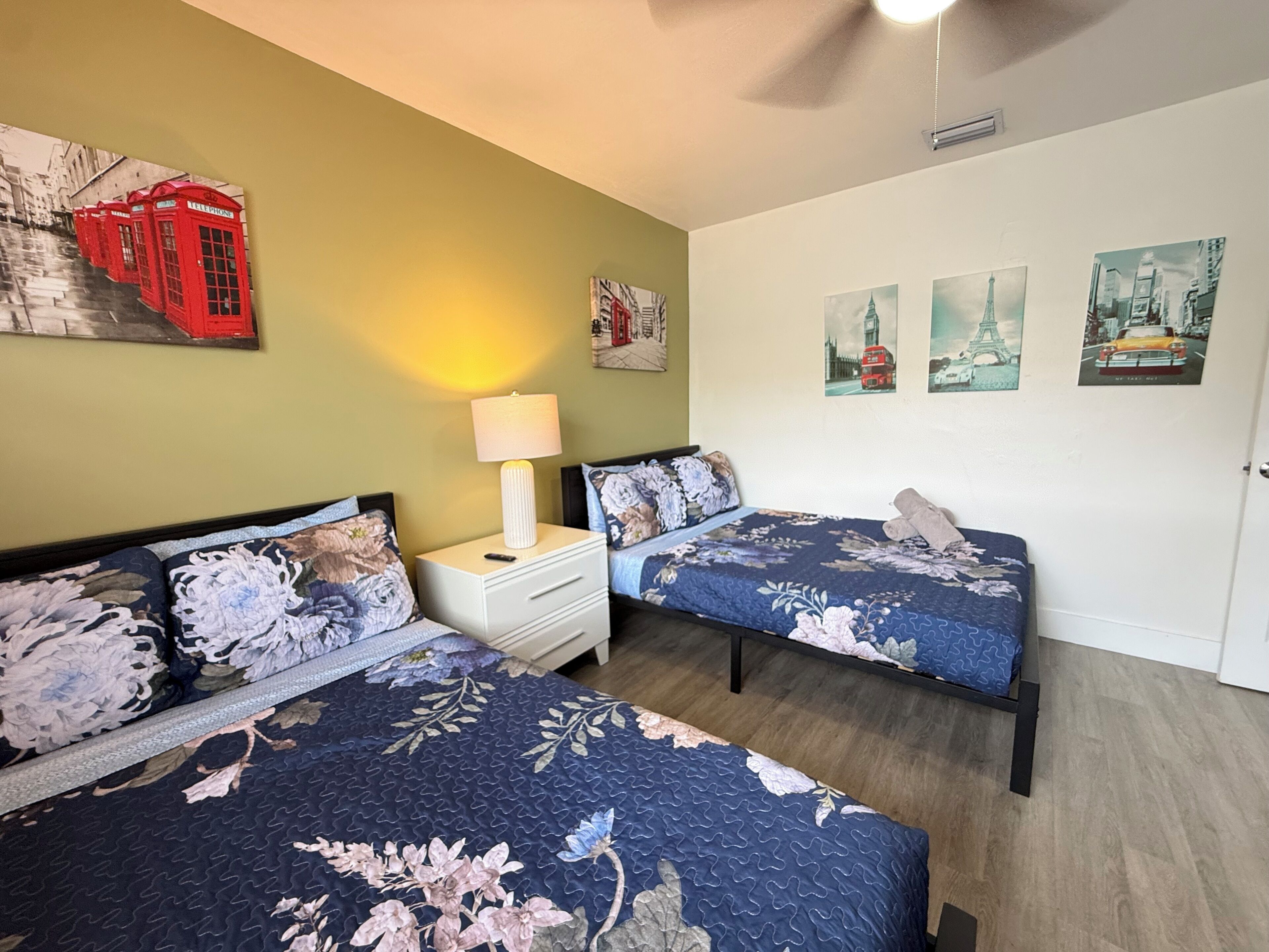 A room with two beds, a nightstand with a lamp, and wall art including a red telephone booth and cityscape pictures.