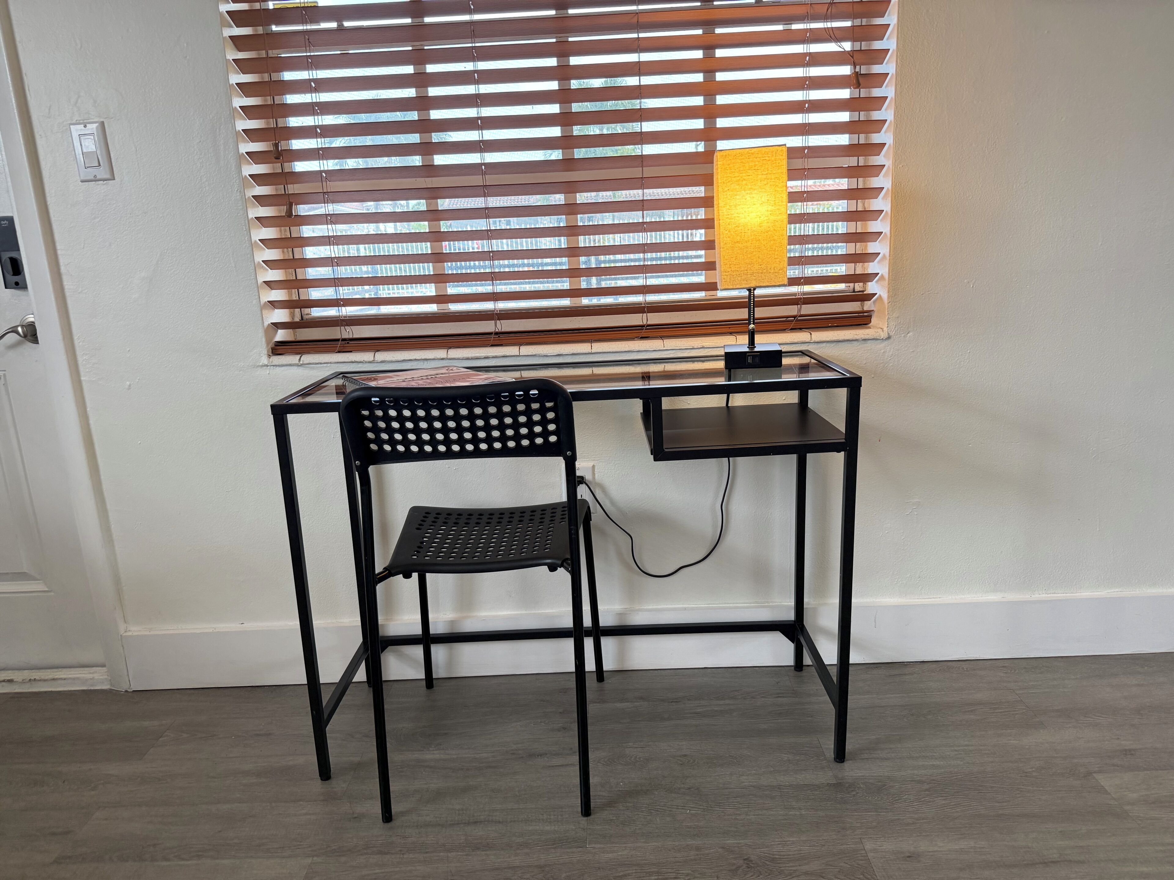 A small black metal desk with a chair, a lamp, and a window with blinds.