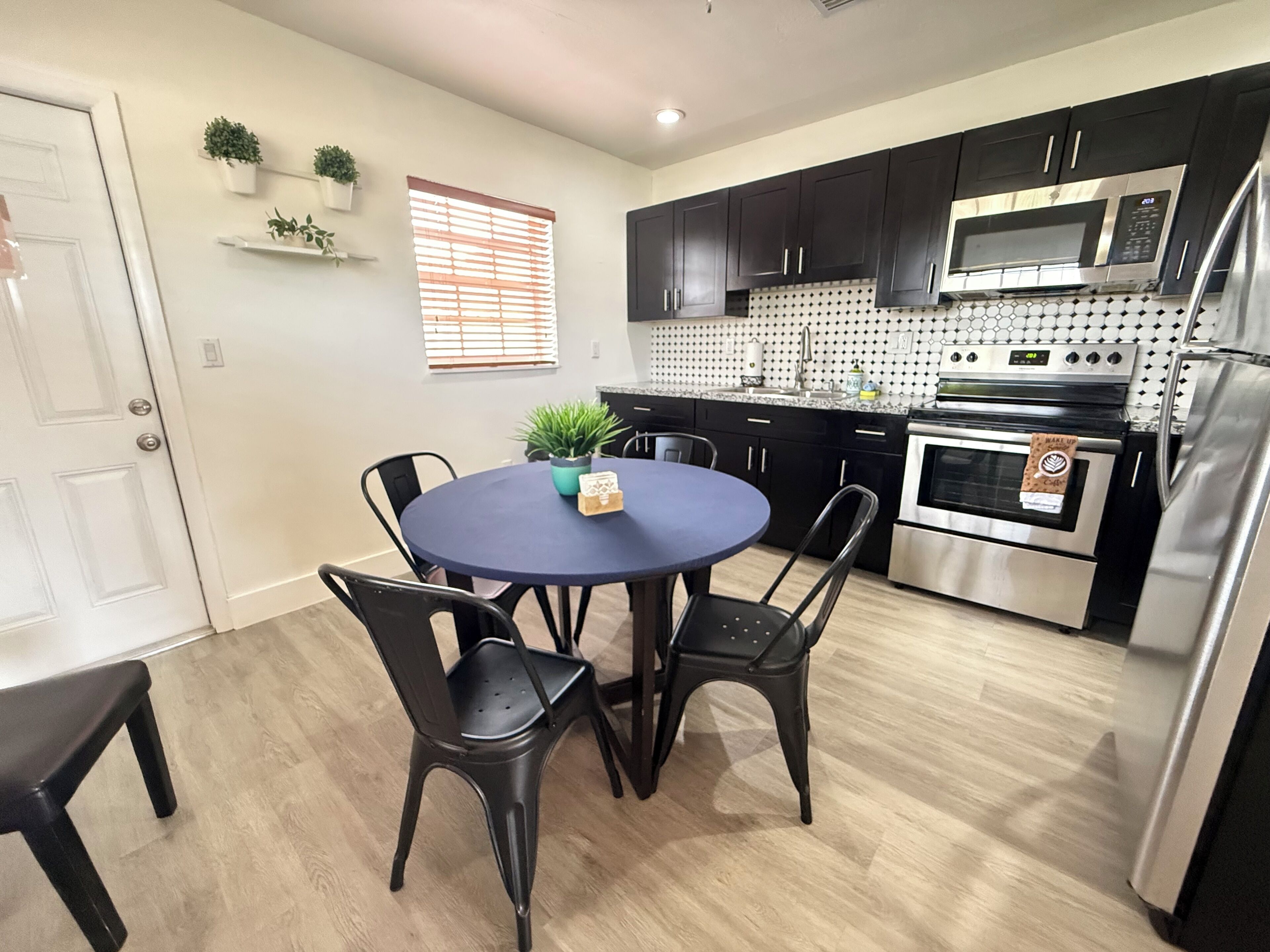 A modern kitchen with black cabinets, a stainless steel refrigerator, a microwave, and an oven. There is a round table with chairs and a potted plant on the table.