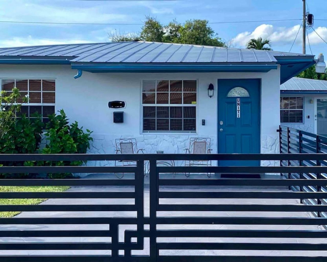 Un edificio a un piano con una porta blu e finestre, un piccolo portico con sedie e un recinto metallico in primo piano.