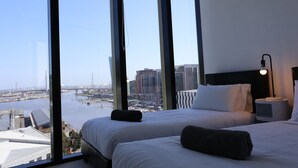 A hotel room with a large window offering a city view, a bed with white linens, a nightstand, and a lamp.