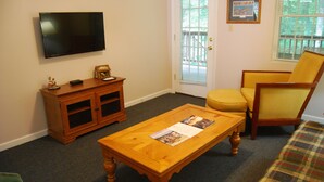 Una sala de estar con televisor de pantalla plana, mueble de madera, sillón amarillo, sofá a cuadros y una mesa de centro con revistas.
