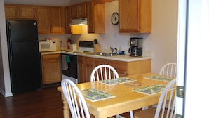Una cocina con armarios de madera, un refrigerador negro, un microondas y una cafetera. Hay una mesa de comedor de madera con sillas y manteles individuales.