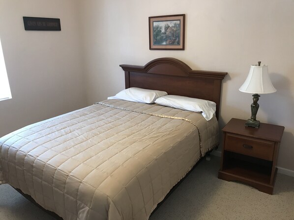 Una cama bien tendida con un edredón color crema, un cabecero de madera, una lámpara de noche y una mesita de noche de madera con un cajón.