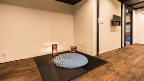 Une pièce avec un plancher en bois, un tapis noir, un coussin à motifs, un petit tabouret et une étagère fixée au mur.