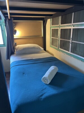 A single bed with a blue mattress and a rolled white towel, a window with a curtain, and a wall with a green trim. - 2Street Hostel at Surathani - Adults Only (Surat Thani)