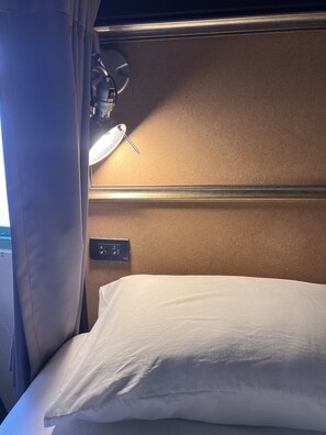 A train compartment with a bed, a headrest, a window with curtains, and a wall-mounted light. - 2Street Hostel at Surathani - Adults Only (Surat Thani)