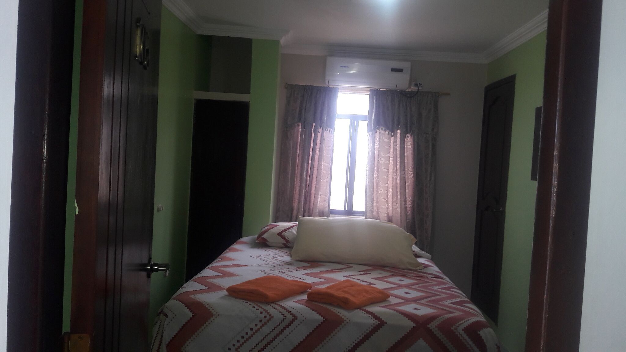 A narrow room with a bed, a window with curtains, and an air conditioning unit.