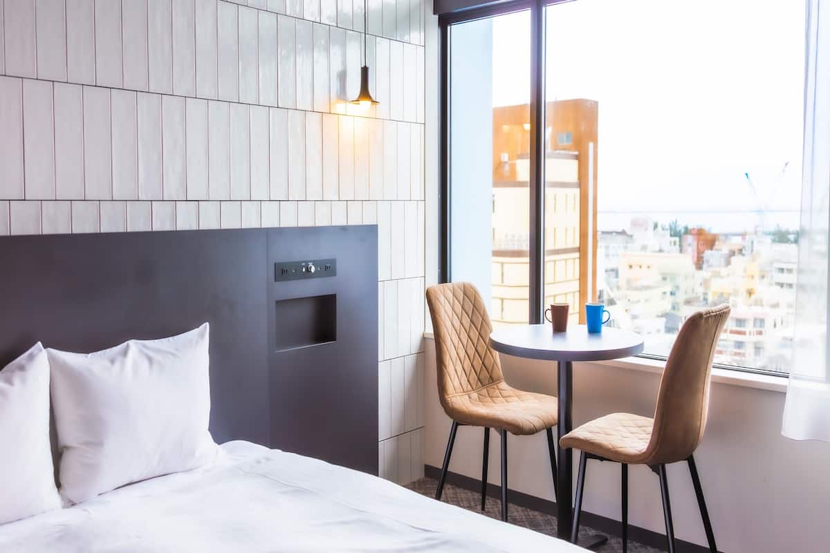 Una habitación de hotel con una cama, una mesita con dos sillas, un ventanal con vistas a edificios y un panel de control en la pared.