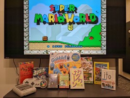 Um jogo de Super Mario World numa televisão, com vários livros de videojogos e manga numa estante abaixo.