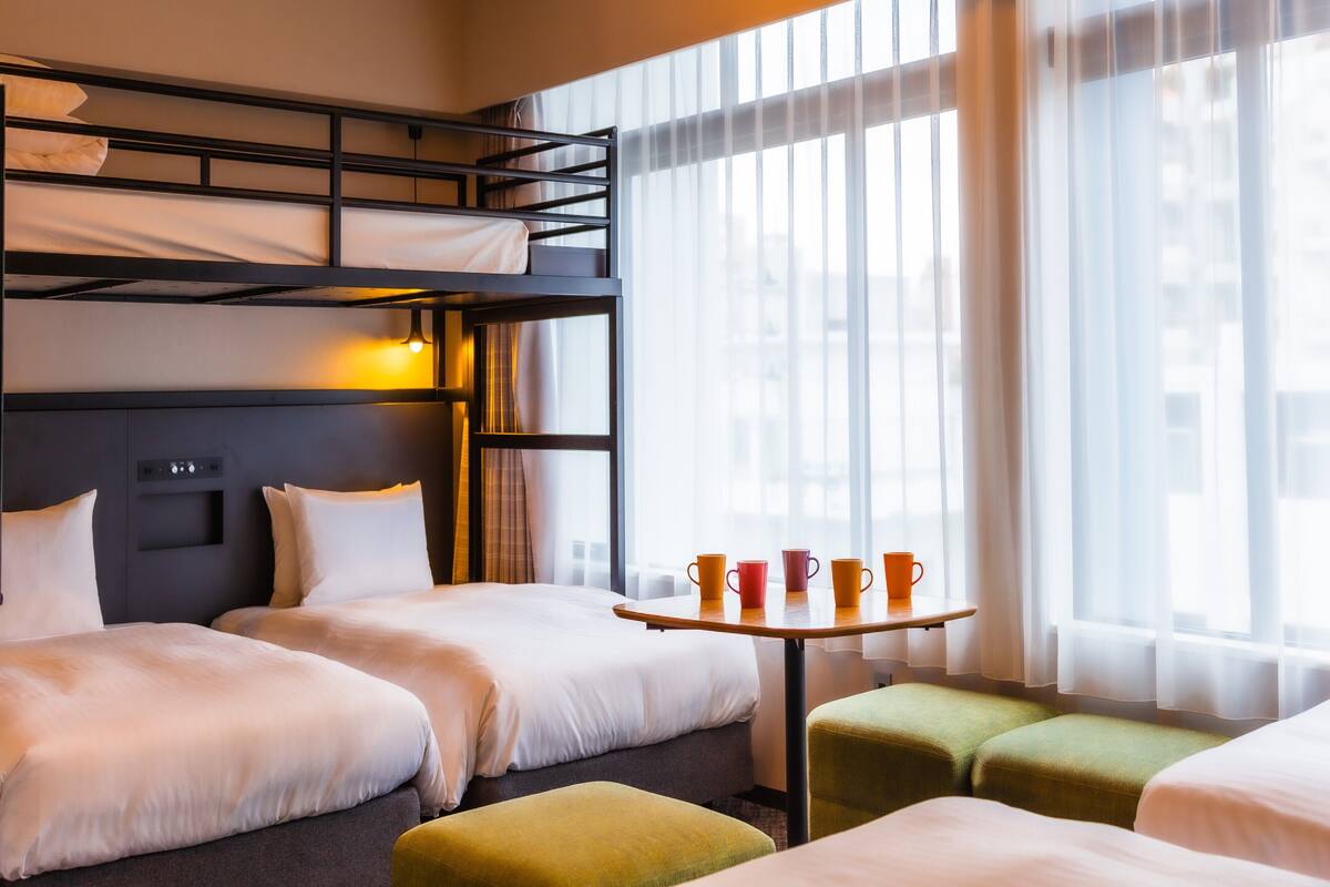 Habitación de hotel con literas, una cama individual, una mesita con tazas y una ventana con cortinas translúcidas.