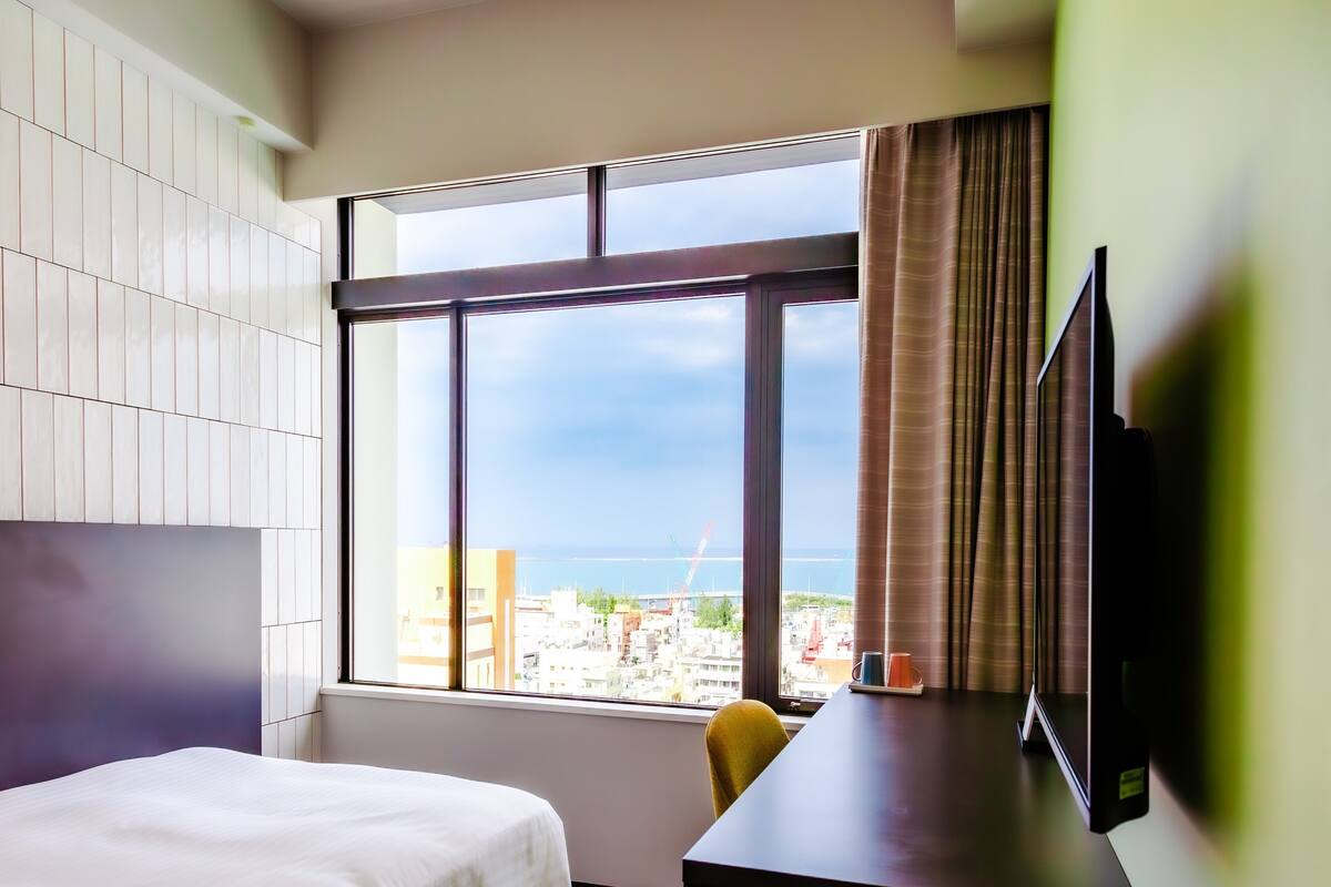 Habitación de hotel con cama, escritorio, televisión y un amplio ventanal con vistas a la ciudad.