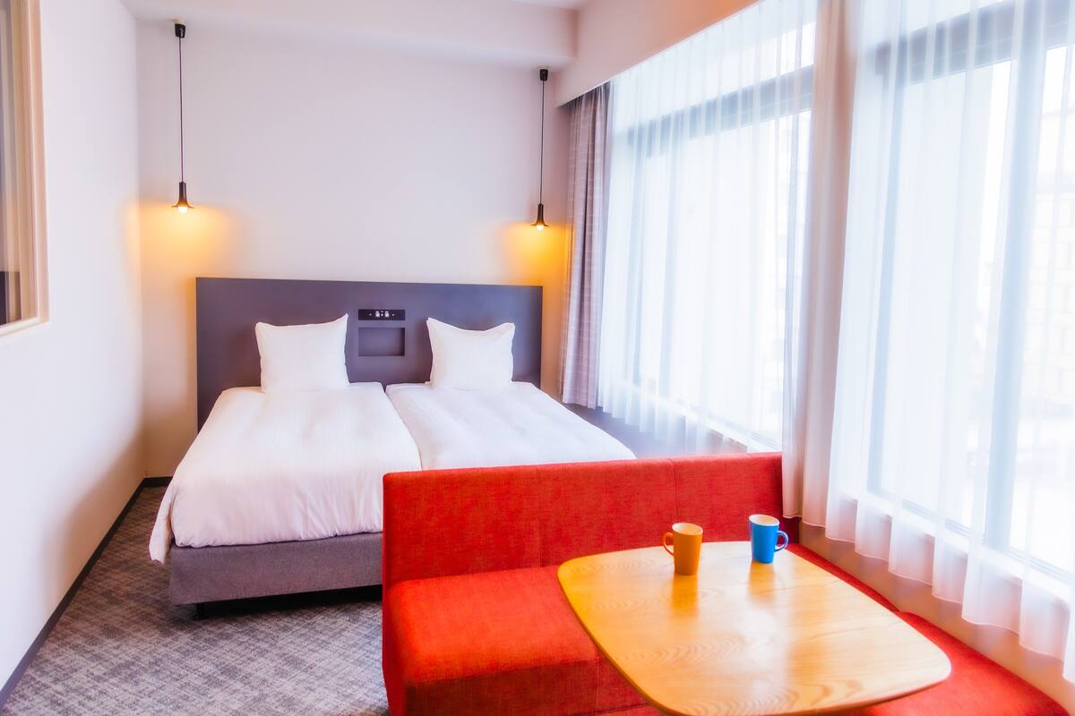 Habitación de hotel con una cama doble, un sofá rojo, una mesita de madera con dos tazas y un ventanal con cortinas de encaje.