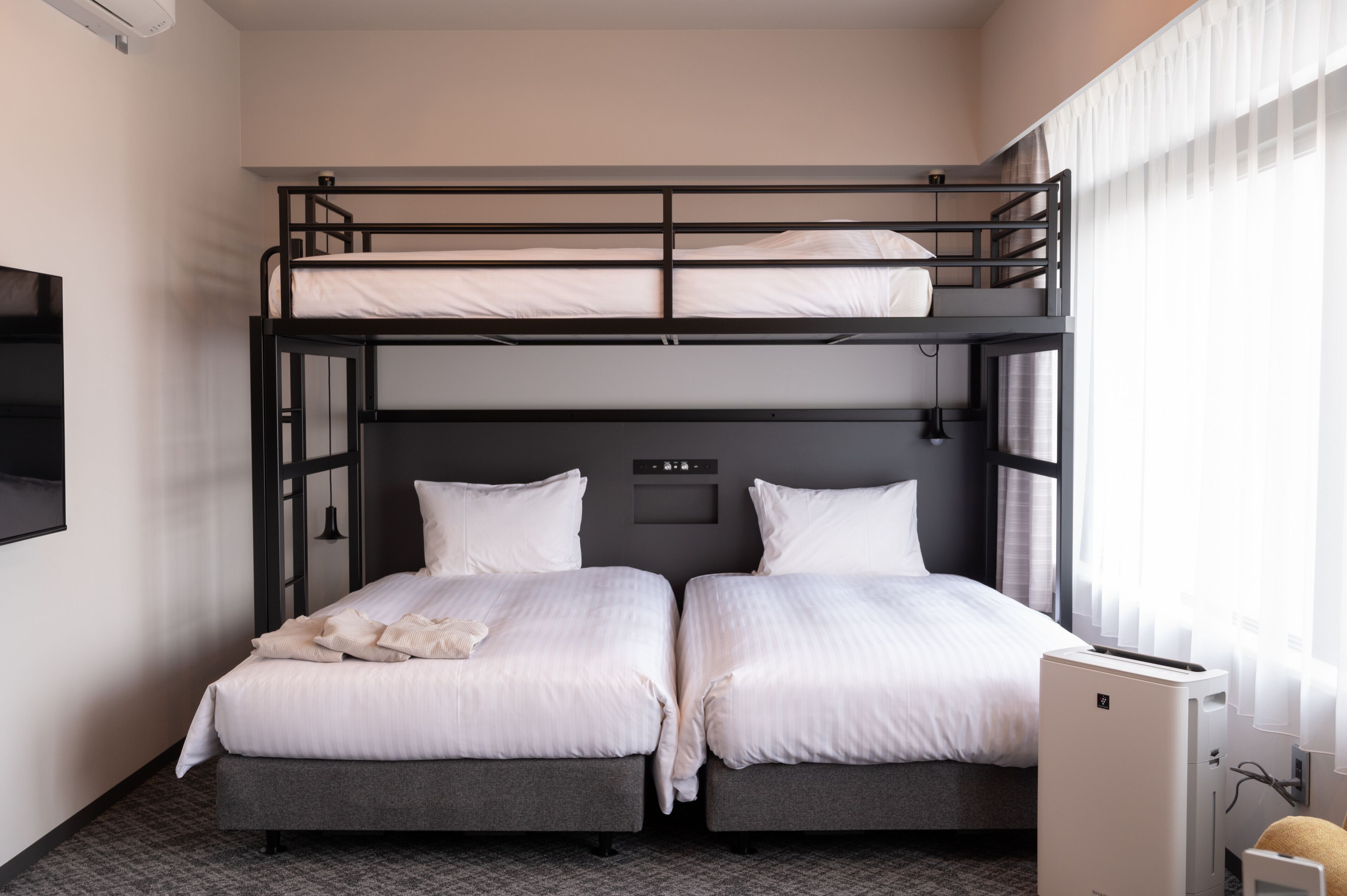 A hotel room with two beds, a bunk bed above, a flat-screen TV, and a small desk with a lamp.