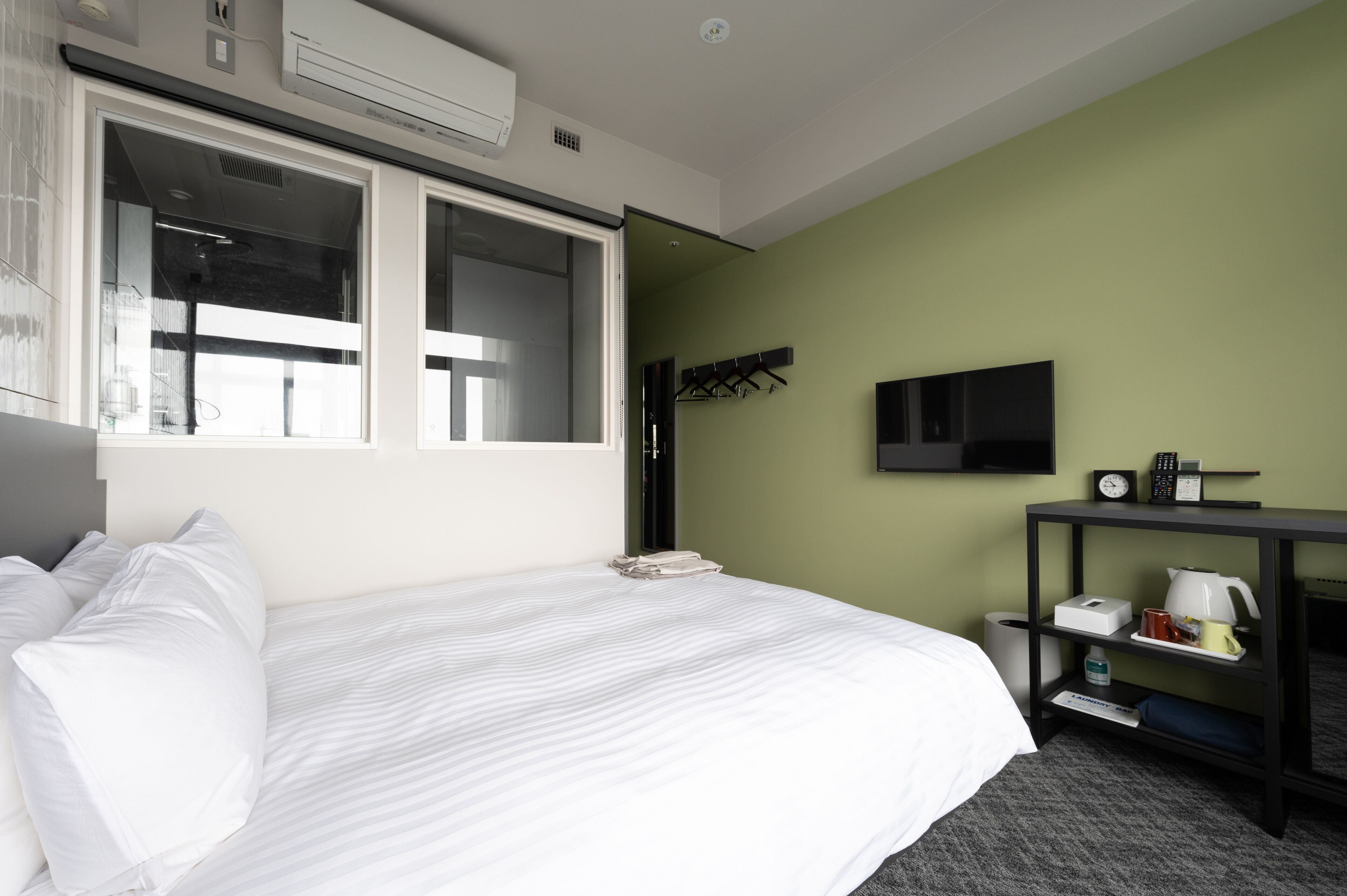 A hotel room with a large bed, a TV mounted on the wall, a small table with a kettle and cups, and a window with a view of the outside.