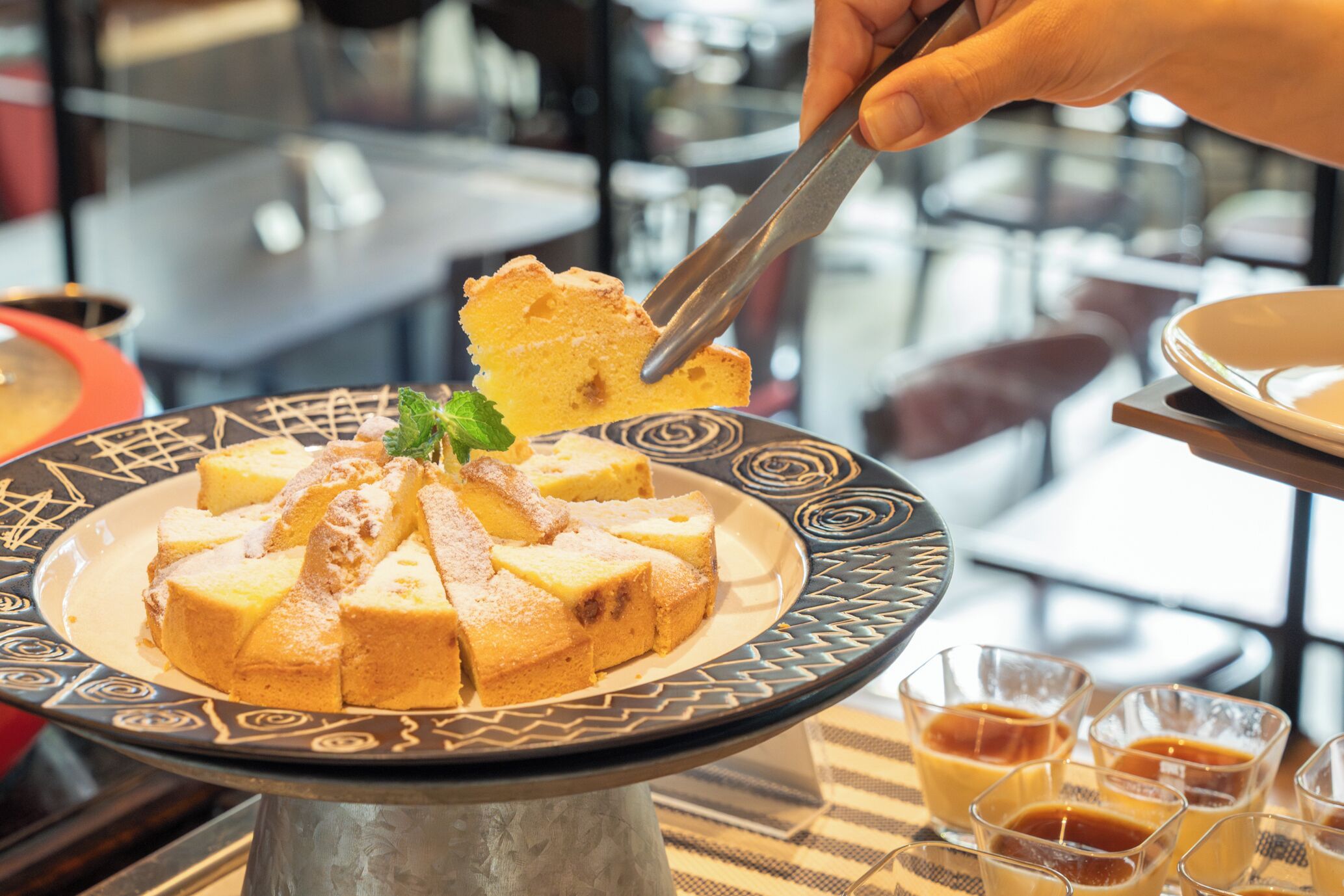 A dessert plate with a cake on a decorative stand, accompanied by small glasses of a dark beverage.