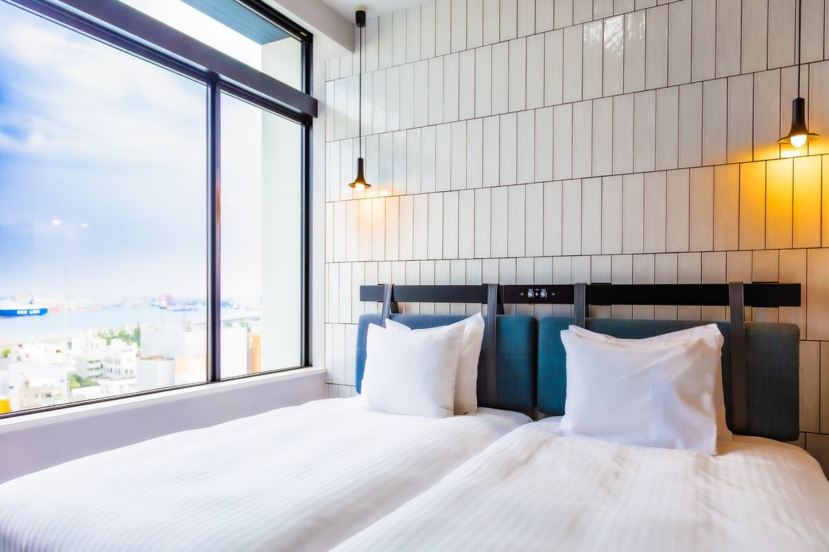 Habitación de hotel con un ventanal grande, cama con ropa blanca y cabecera con panel de control.