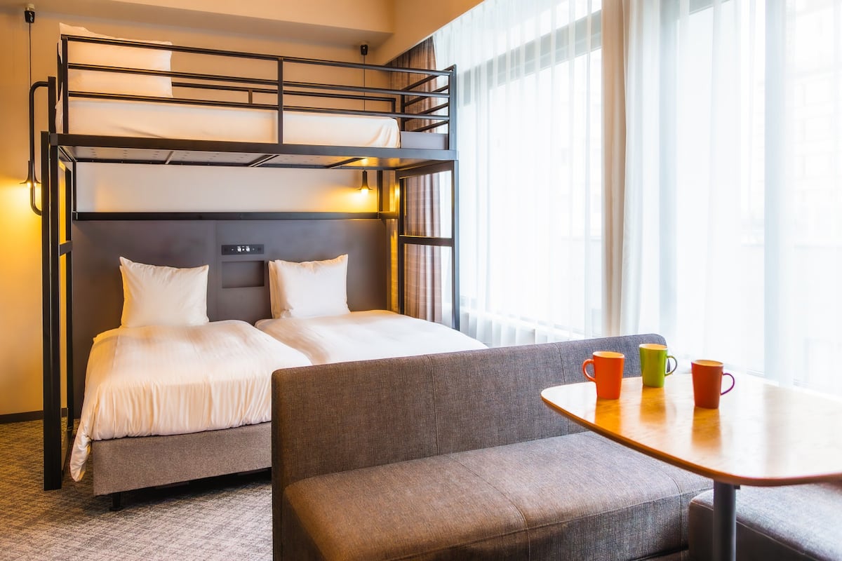 Habitación de hotel con literas, un sofá, una mesita pequeña con tazas de colores y una ventana con cortinas translúcidas.