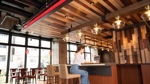 A woman seated at a bar with a laptop, in a modern interior with wooden beams, large windows, and hanging pendant lights. - Hotel Cocktail Stay Naha (Naha)