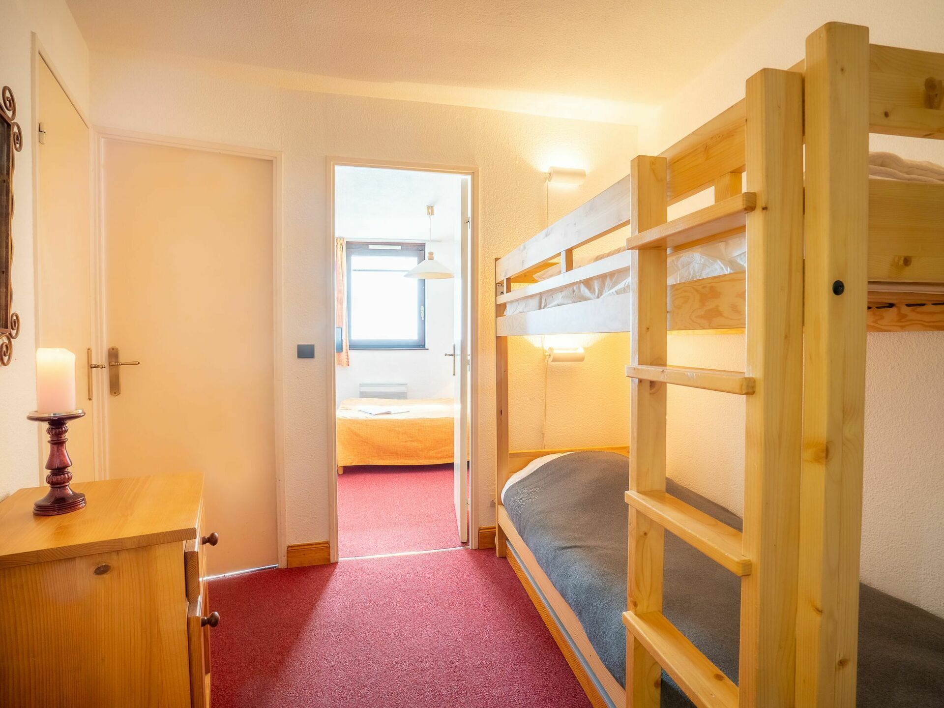 Een kamer met een stapelbed, een houten dressoir, een deur naar een andere kamer en een rood tapijt.