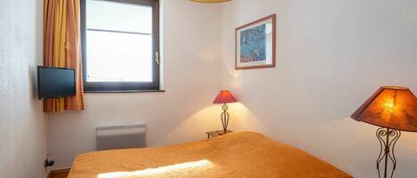 A room with a bed, a window with curtains, a wall-mounted TV, a framed picture, a table lamp, and a remote control on a small table.