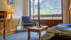 Une pièce chaleureuse avec une grande fenêtre donnant sur les montagnes, une chaise en bois, une petite table avec une bouteille et des verres, et un téléviseur posé sur un meuble en bois.
