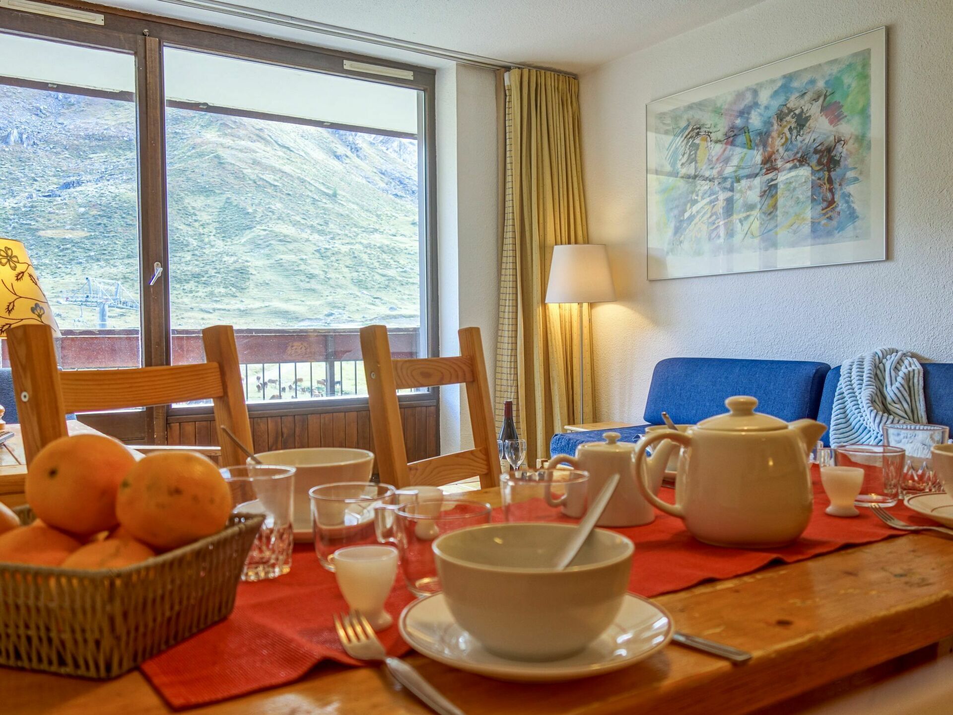 Une table à manger avec des aliments pour le petit-déjeuner, un panier d’oranges et une vue sur un paysage montagneux par la fenêtre.