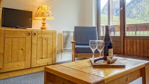 Une pièce chaleureuse avec un meuble en bois, une télévision, une lampe, une chaise, une table avec des verres à vin et une bouteille, et une fenêtre donnant sur les montagnes.