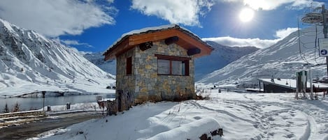 Une cabane en pierre avec un toit en bois, entourée de montagnes enneigées et surplombant un ciel dégagé.
