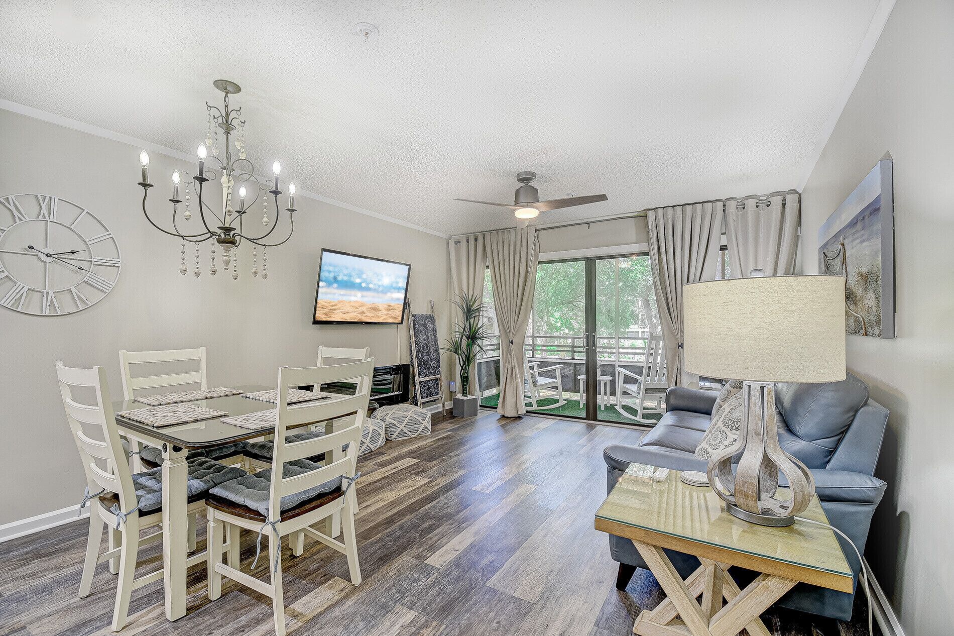 A living room with a dining area, a ceiling fan, a TV, and a balcony with a view of greenery.
