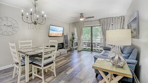En stue med spiseområde, loftventilator, fjernsyn og en balkon med udsigt over grønne omgivelser.