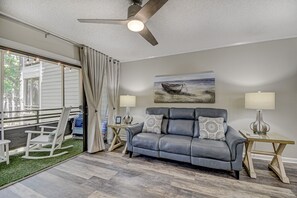 A living room with a gray leather sofa, two side tables with lamps, a ceiling fan, a painting of a boat on the wall, and a view of the outdoors through a sliding glass door.
