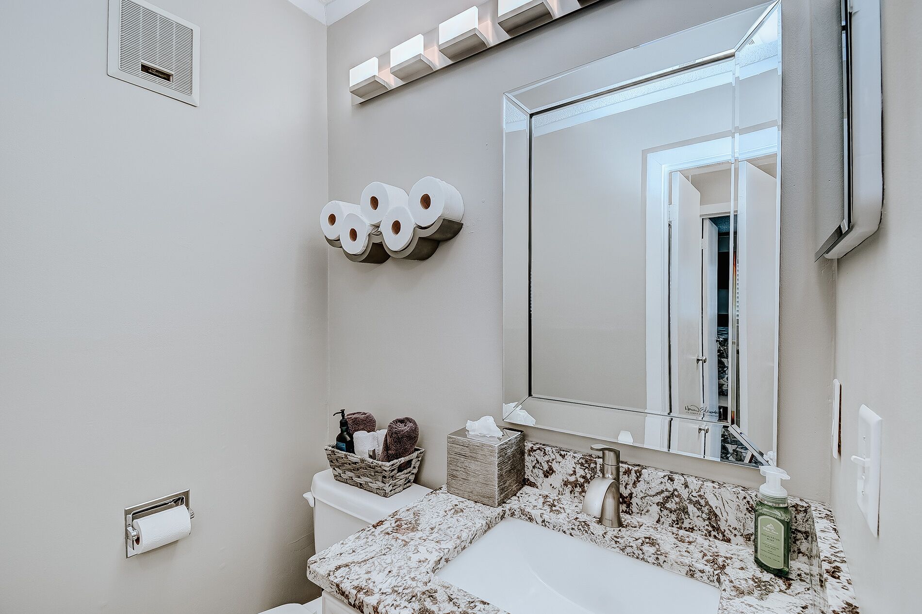 A bathroom with a toilet paper holder, a mirror, and a sink with a marble countertop.