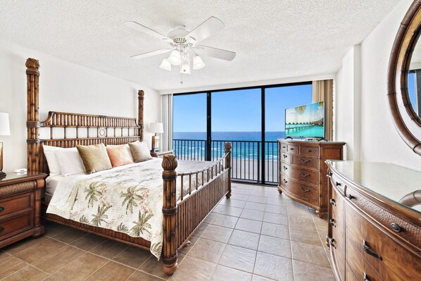 A bedroom with a large bed, wooden headboard, a ceiling fan, a nightstand, a dresser, and a large window with a view of the ocean.