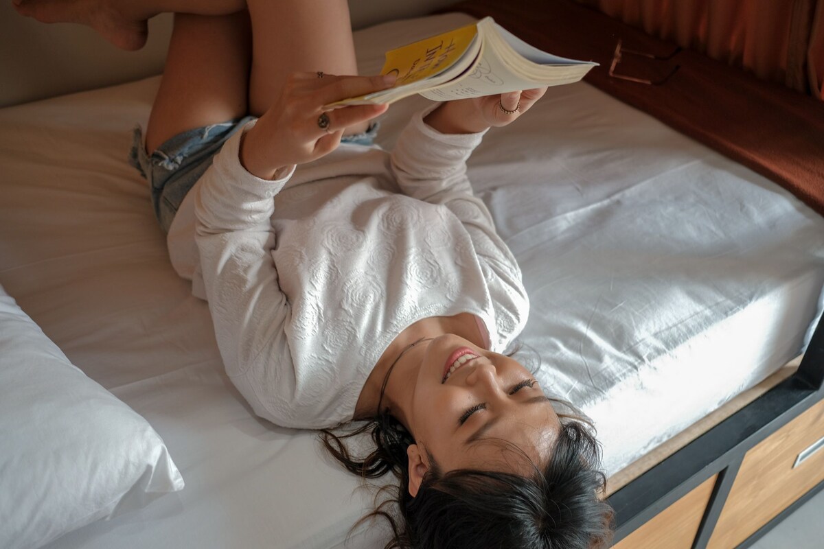 A person lying on a bed with a book, in a room with a wooden headboard and a window.