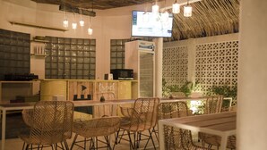 A restaurant with wicker chairs, a long table, and a TV mounted on the wall. - Canggu Castle - Hostel (Tibubeneng)