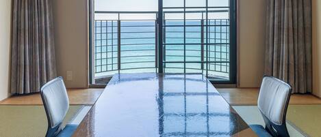 A room with a large table, two chairs, a view of the sea through a window with a metal grid, and traditional tatami mats on the floor.