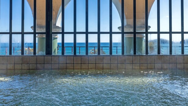 A view through a window with a tiled floor and a railing, overlooking a body of water and a structure with arches. - Hotel Riviera Shishikui (Kaiyo)