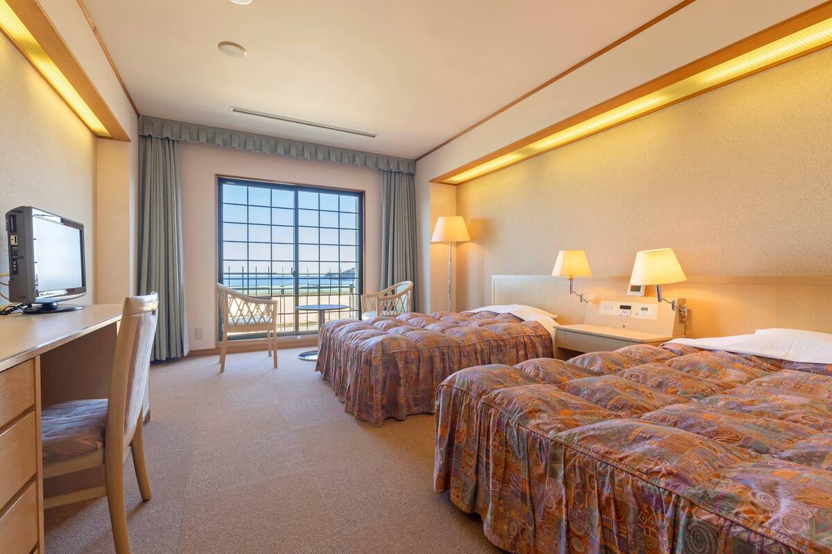 A hotel room with two beds, a desk with a computer, a chair, and a window with a view.