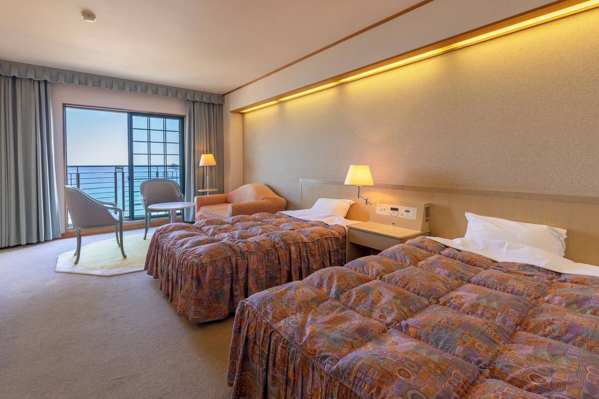 A hotel room with two beds, a chair, a nightstand, and a large window with curtains.