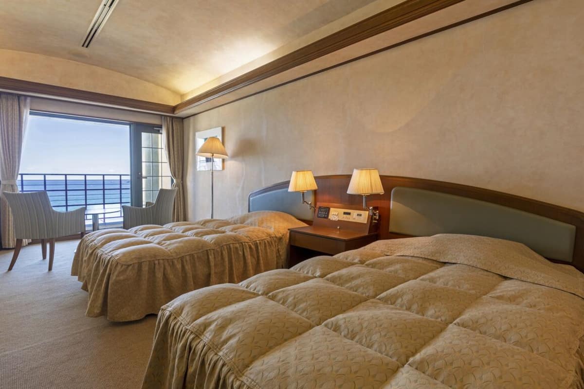 A hotel room with two beds, a desk with lamps, a chair, and a large window with curtains.