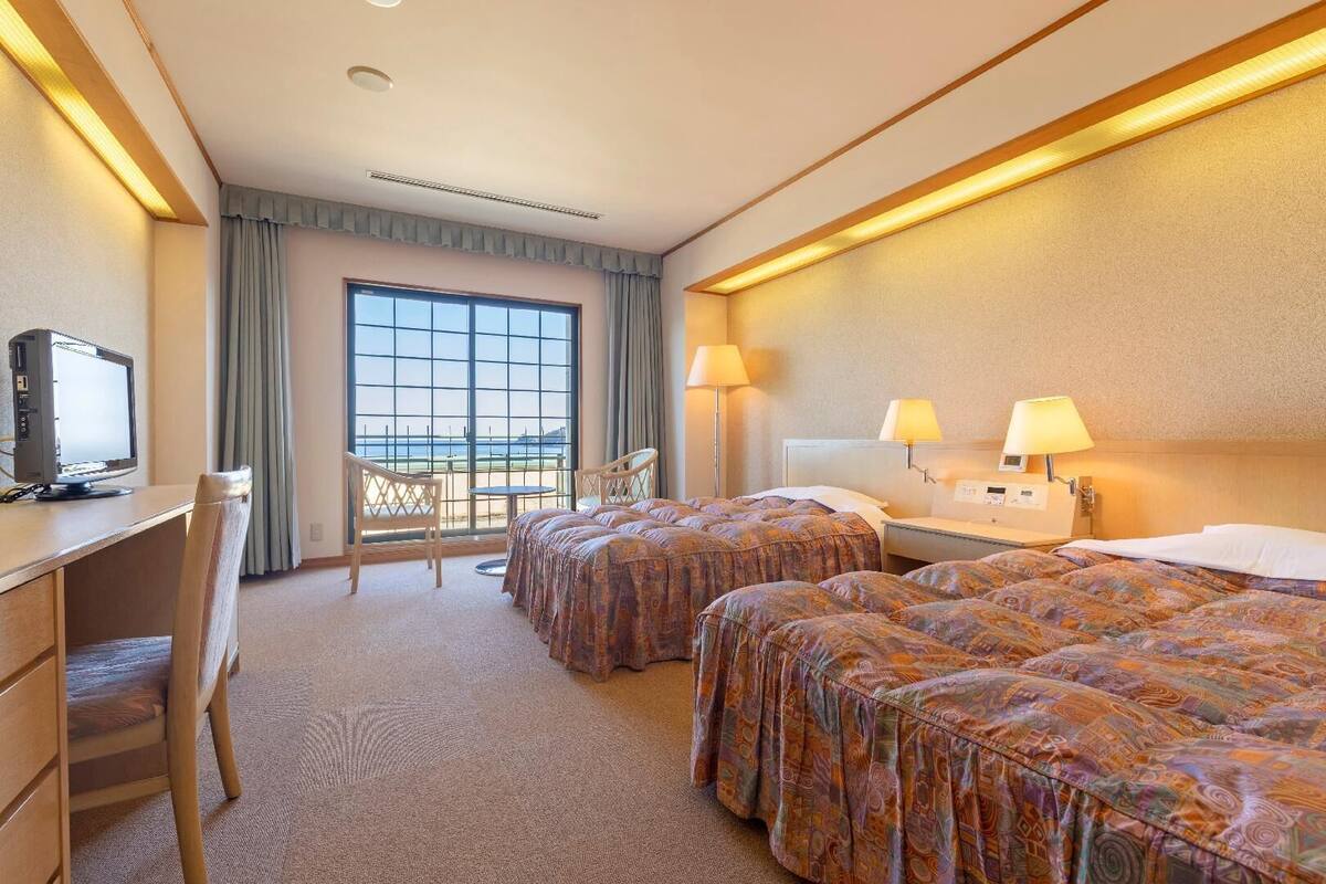 A hotel room with two beds, a desk with a computer, a chair, and a view of the sea through large windows.