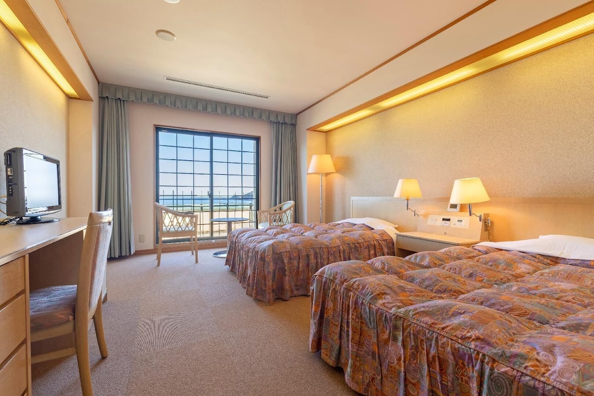A hotel room with two beds, a desk with a computer, a chair, and a view of the sea through large windows.